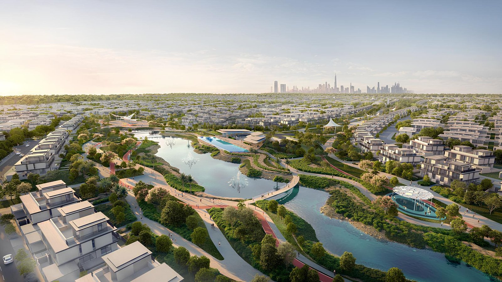 Aerial view of a landscaped master community — townhouses and low-rise apartment buildings set around a central lagoon and parkland, with the Dubai skyline on the horizon